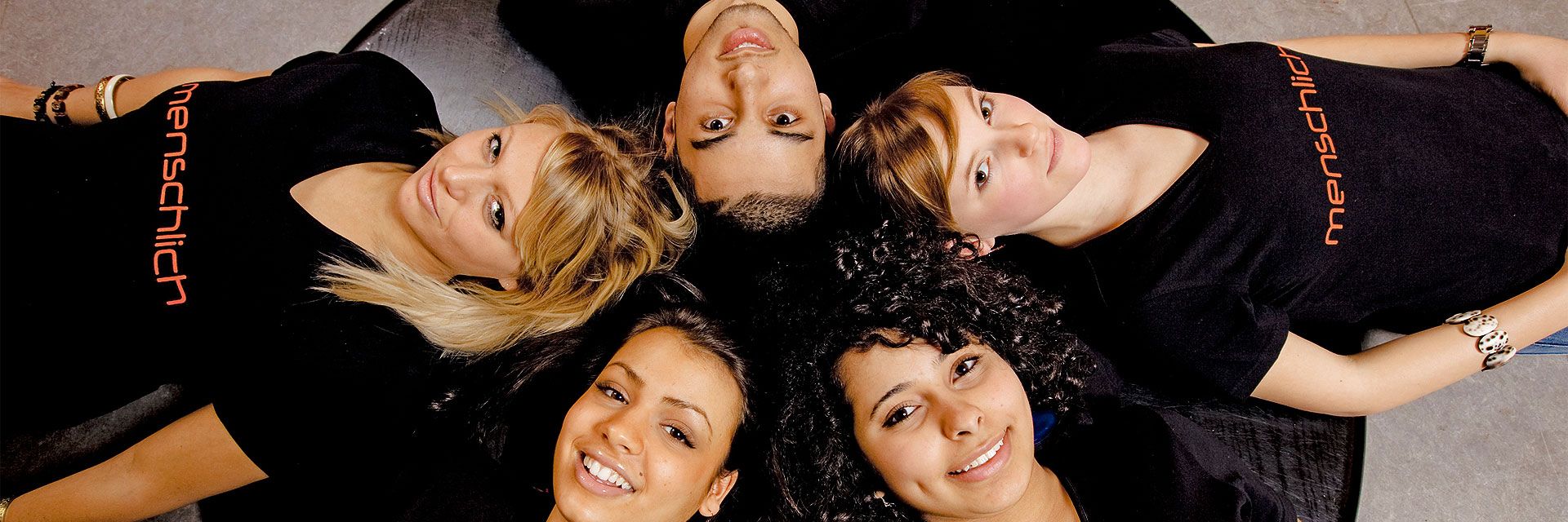 Foto: Fünf Jugendliche liegen Kopf an Kopf im Kreis. Sie lachen und wirken vergnügt und herzlich. Alle tragen ein schwarzes T-Shirt mit der Aufschrift 'menschlich'.
