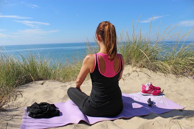 Foto: In einer Düne nahe des Meeres sitzt eine junge Frau gelassen auf ihrer Yogamatte. Sie kommt zur Ruhe ohne einzuschlafen.