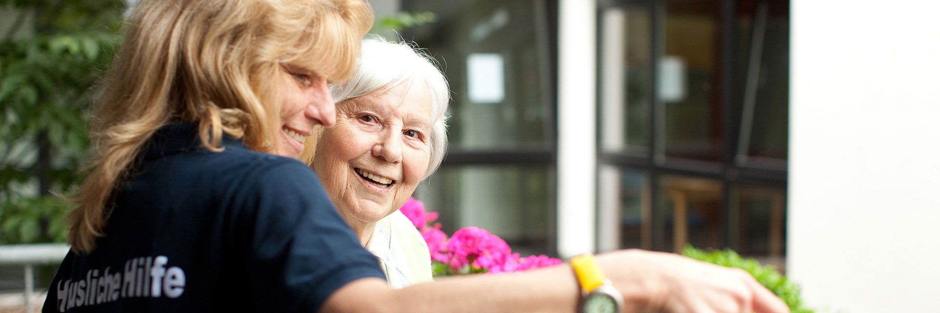 Foto: Eine DRK-Mitarbeiterin gießt die Balkonblumen in der Wohnung einer älteren Dame. Die ältere Dame wirkt glücklich und konzentriert sich mehr auf das gemeinsame Gespräch als auf die Blumen.